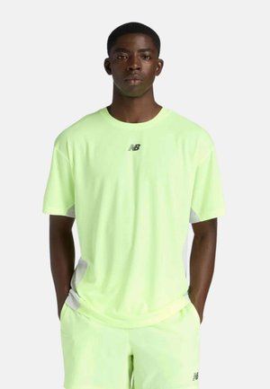 Joven con las manos en los bolsillos, vestido con una camiseta deportiva New Balance verde clara y pantalones cortos a juego, sobre un fondo liso.