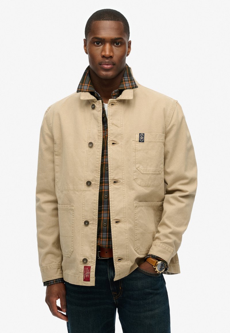 Beige Canvasjacke mit einer Knopfleiste, zwei Taschen und einem karierter Flanellshirt darunter. Dunkle Jeans und eine Armbanduhr sind ebenfalls sichtbar.