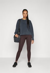 Felpa grigia scura, leggings marroni a coste e sneakers nere. Una borsa a tracolla nera è tenuta nella mano sinistra. Design semplice.