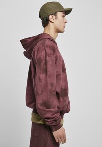 Hanorac tie-dye în nuanța bordo, cu buzunar tip kangaroo, glugă cu șnur și manșete ribbate. Purtați peste un top verde și cu pantaloni asortați.