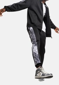 Mann in schwarzen Jogginghosen mit weißen und grauen Schlangenmuster-Seitenstreifen, passenden Sneakers, einer schwarzen Jacke und einem weißen Shirt, im Schritt.