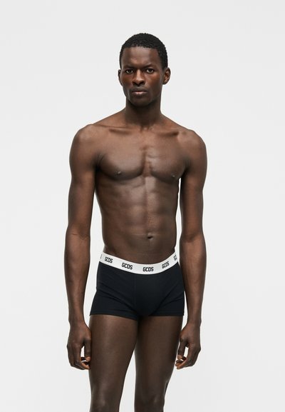 Αθλητικός άνδρας με σκούρο δέρμα και κοντά μαλλιά, φοράει μαύρα μπόξερ της GCDS, στέκεται μπροστά σε απλό ανοιχτό γκρι φόντο.