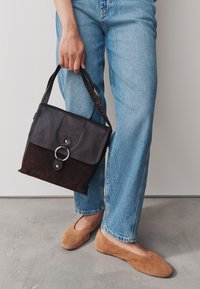 Geantă de mână din piele și piele întoarsă maro închis, cu clapetă frontală, accesorii din inel argintiu și curea ajustabilă, ținută alături de blugi albastru deschis.