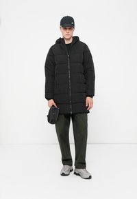 Black puffer coat with horizontal quilted design, zip closure, attached hood, paired with olive-green pants and grey sneakers.