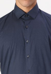 Navy button-up shirt featuring a geometric pattern of small circles, with a structured collar and matching dark buttons. Soft fabric texture.