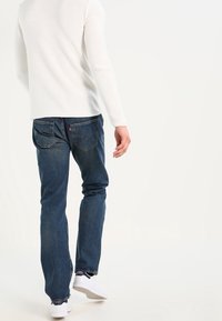 Jeans denim biru dengan potongan lurus, dilengkapi saku belakang dan hem yang kasar. Dipadukan dengan kemeja lengan panjang tekstur putih dan sepatu sneakers putih.