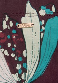 Donker bordeauxkleurige stof met een bloemenpatroon met witte en teal bloemblaadjes, afgewisseld met stippen; inclusief een goudkleurig merklabel.