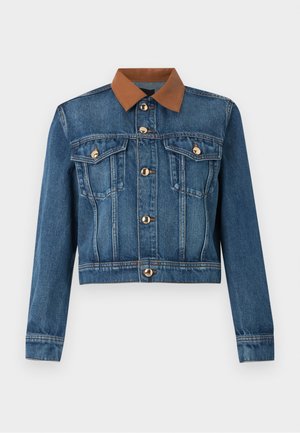 Model draagt een spijkerjack met een bruine kraag, gestreept paarse polo, beige korte broek, bruine sokken en bruine plateauzolen, staat met handen in de zakken.