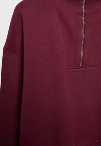 Sweat-shirt bordeaux à manches longues avec une fermeture éclair partielle à l'avant et une texture côtelée autour du col et de la zone de la fermeture éclair.