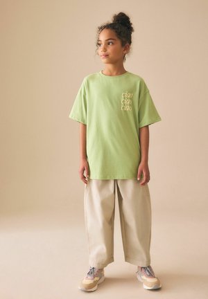 Fiatal, göndör hajjal, kontyban, zöld, túlméretezett "ciao" feliratú pólót, bézs bő szárú nadrágot és színes tornacipőt viselve, mozdulatlanul áll.