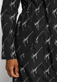 Czarna kurtka z białym, powtarzającym się wzorem napisu "Hugo", przednia kieszeń z zapięciem na zatrzask i ręka spoczywająca na boku.