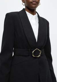 Μαύρο σακάκι με γιακά σάλι, με ζώνη στη μέση με χρυσή οβάλ αγκράφα. Διαθέτει λευκό πουκάμισο με υφή από μέσα και πλαϊνές τσέπες.