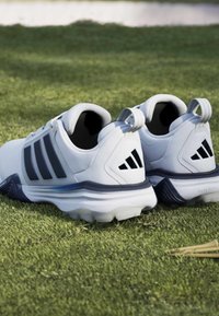 Άσπρα και μαύρα αθλητικά παπούτσια Adidas τοποθετημένα πάνω σε πράσινο γρασίδι με tees του γκολφ κοντά.