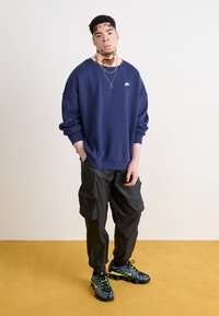 Námořnický modrý oversize svetr s bílým logem, spárovaný s černými cargo kalhotami s bočními kapsami a černými a neonově zelenými sportovními botami.
