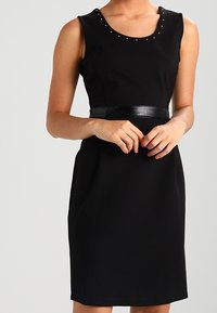 Kobieta w bezrękawowej, czarnej sukience do kolan z kolnierzykiem ozdobionym ćwiekami i błyszczącym czarnym paskiem, z rękami złożonymi na przodzie.