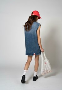 Denim ujjatlan ruha fokozatos színátmenettel, piros sapkával és fekete platform cipőkkel párosítva. Egy fehér mintás vászontáskát tart.