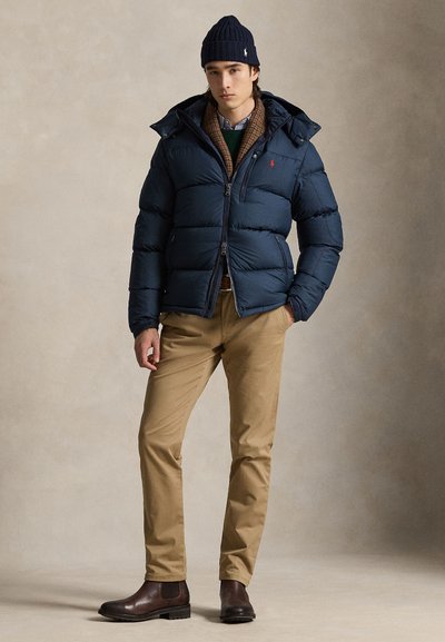 Ung mand iført navy dunjakke, navy hue, beige bukser og brune støvler, stående med den ene hånd i lommen mod neutral baggrund.