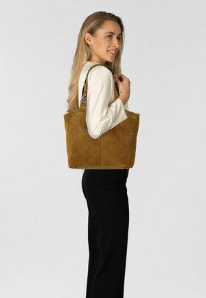 Sac fourre-tout en daim marron avec un design spacieux, doté de deux bandoulières et de détails en métal argenté, présenté sur un fond neutre.