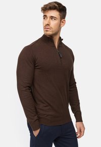 Suéter marrón de manga larga con cremallera y cuello alto, puños acanalados y textura suave. Presenta un detalle de tira de cremallera en contraste.
