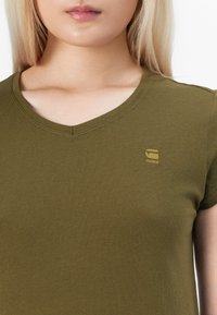 Alyvuogių spalvos medvilnės marškinėliai su V formos iškirpte, trumpomis rankovėmis ir mažu įspaustu logotipu šviesesnėje spalvoje krūtinės srityje.
