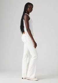 Hvite utsvingte jeans laget av denim, med kontraststing og en brun lærlapp. Kombinert med en hvit tank-top og joggesko.