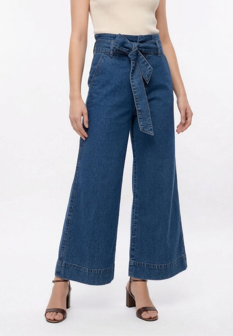 Γυναίκα που φοράει μπλε τζιν παντελόνια με υψηλή μέση και φαρδύ πόδι, με μπροστινή ζώνη που δένει, συνδυασμένα με καφέ πέδιλα με τακούνι και μπεζ αμάνικη μπλούζα.