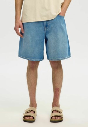 Homme portant un short en jean bleu clair, des sandales beiges et une chemise beige, une main dans la poche, debout sur fond blanc.