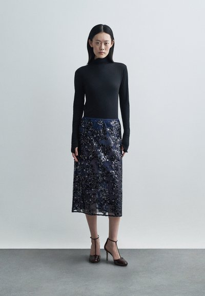 Μαύρη μπλούζα με μακρύ μανίκι, συνδυασμένη με διαφανή ναυτική φούστα με λουλουδάτα μοτίβα και παγιέτες. Καφέ γόβες με λουράκι στον αστράγαλο συμπληρώνουν την εμφάνιση.