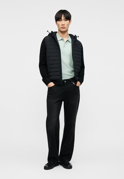 Νέος άνδρας που φοράει μαύρο καπιτονέ μπουφάν με κουκούλα, ανοιχτό πράσινο πόλο πουκάμισο, μαύρο τζιν ευθείας γραμμής και μαύρα αθλητικά παπούτσια, στέκεται με τα χέρια στις τσέπες.