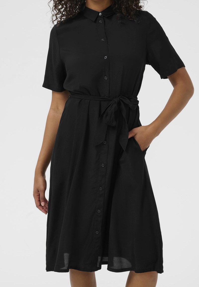 Femme portant une robe noire boutonnée au genou, manches courtes et ceinture nouée à la taille, une main dans la poche.