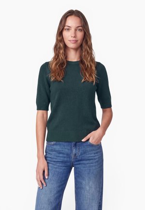 Femme aux cheveux bruns ondulés portant un pull manches courtes vert foncé et un jean bleu, debout avec une main dans sa poche.