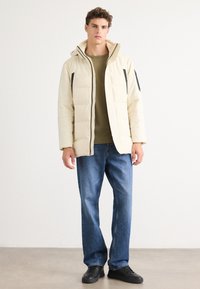 Beige Daunenjacke mit Kapuze, versehen mit schwarzen Akzenten, Reißverschluss und überlappenden Taschen. Trägt man über einem olivgrünen Oberteil und blauen Jeans.