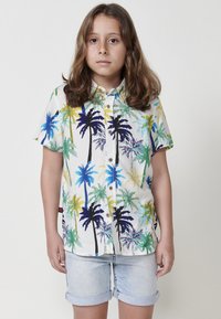 Camisa de botones de manga corta, con un vívido estampado de palmeras en azul, verde, amarillo y negro sobre un fondo blanco. Llevada con pantalones cortos de mezclilla.