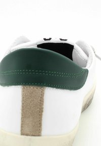 Sneaker bianco con un tallone in pelle verde e un dettaglio in suede color beige. Presenta accenti di cucitura. Suola in gomma visibile sul bordo inferiore.