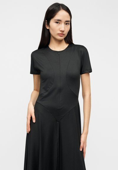 Μαύρο φόρεμα από λείο ύφασμα, με εφαρμοστή κορυφή, κοντά μανίκια και φερμουάρ στο ντεκολτέ· ανοίγει απαλά από τη μέση.