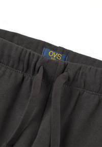Donkergrijze katoenen joggers met een elastische tailleband, trekkoord en een blauwe label met "OVS kids" in het geel. Zachte textuur en casual ontwerp.