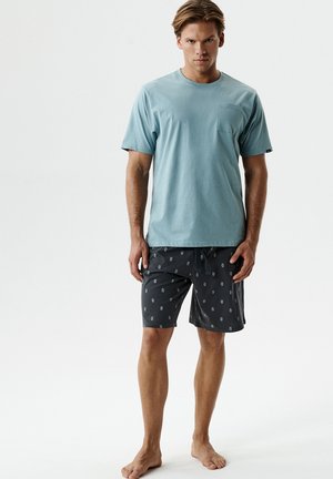 Młody mężczyzna stoi boso, ubrany w jasnoniebieską koszulę z krótkim rękawem i ciemne, wzorzyste spodenki na jednolitym białym tle.