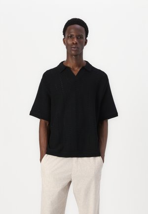 Polo shirt nero in maglia con maniche corte, caratterizzato da un motivo texturizzato e un design a V. Abbinato a pantaloni beige chiaro.