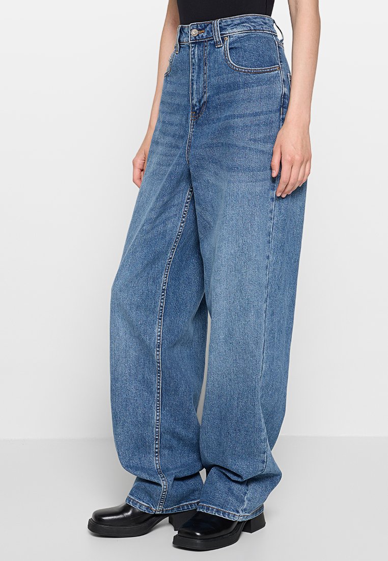 Jean en denim bleu taille haute, jambes larges, avec un délavage, comportant des poches avant et une fermeture à bouton. Porté avec des bottes à plateforme noires.