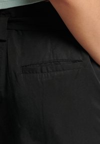Pantaloni in tessuto nero con una piccola tasca posteriore rettangolare, texture liscia e un delicato luccichio del materiale.