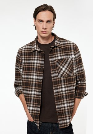 Nuori mies pukeutunut ruskeavalkoiseen ruudulliseen nappipaitaan tummanruskean t-paidan päällä, poseeraa käsineen taskuissa valkoista taustaa vasten.
