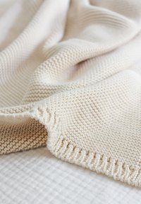 Cream-colored knit blanket with a textured zigzag pattern and braided edge, draped softly over a white quilted surface.