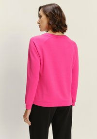 Felpa fucsia con collo a giro, polsini e orlo a coste. Tessuto liscio, vestibilità rilassata e maniche raglan. Pantaloni neri visibili.