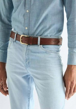 Mann trägt hellblaues Jeanshemd und Jeans mit einem dunkelbraunen Ledergürtel mit goldener Schnalle.