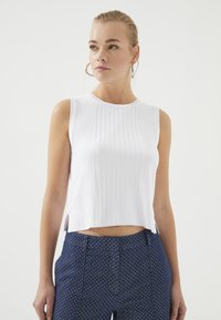 Witte mouwloze top met geribbelde textuur en ronde halslijn, met zijsplitten. Gepaard met marineblauwe broek met witte stippen.