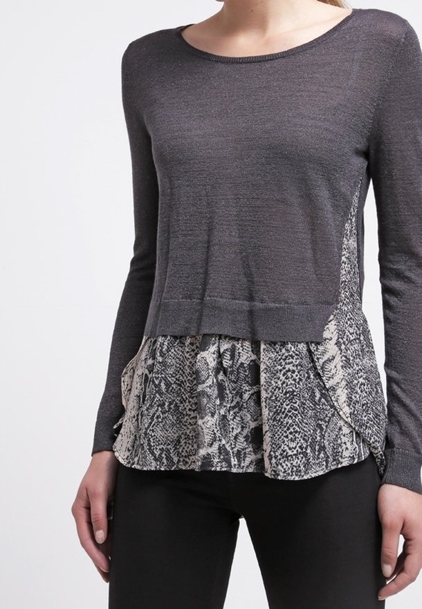 Haut à manches longues gris foncé avec un tissu à imprimé serpent superposé visible sous l'ourlet, porté avec un pantalon noir.