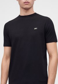 Άνδρας που φοράει απλό μαύρο κοντομάνικο T-shirt με μικρό ορθογώνιο λογότυπο Karl Lagerfeld στο πάνω αριστερό μέρος του στήθους.