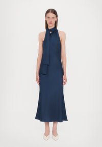 Navy halterneck kjole med bindedetalje, blød silhuet og glat tekstur; har en knælang kant og minimalistisk design.