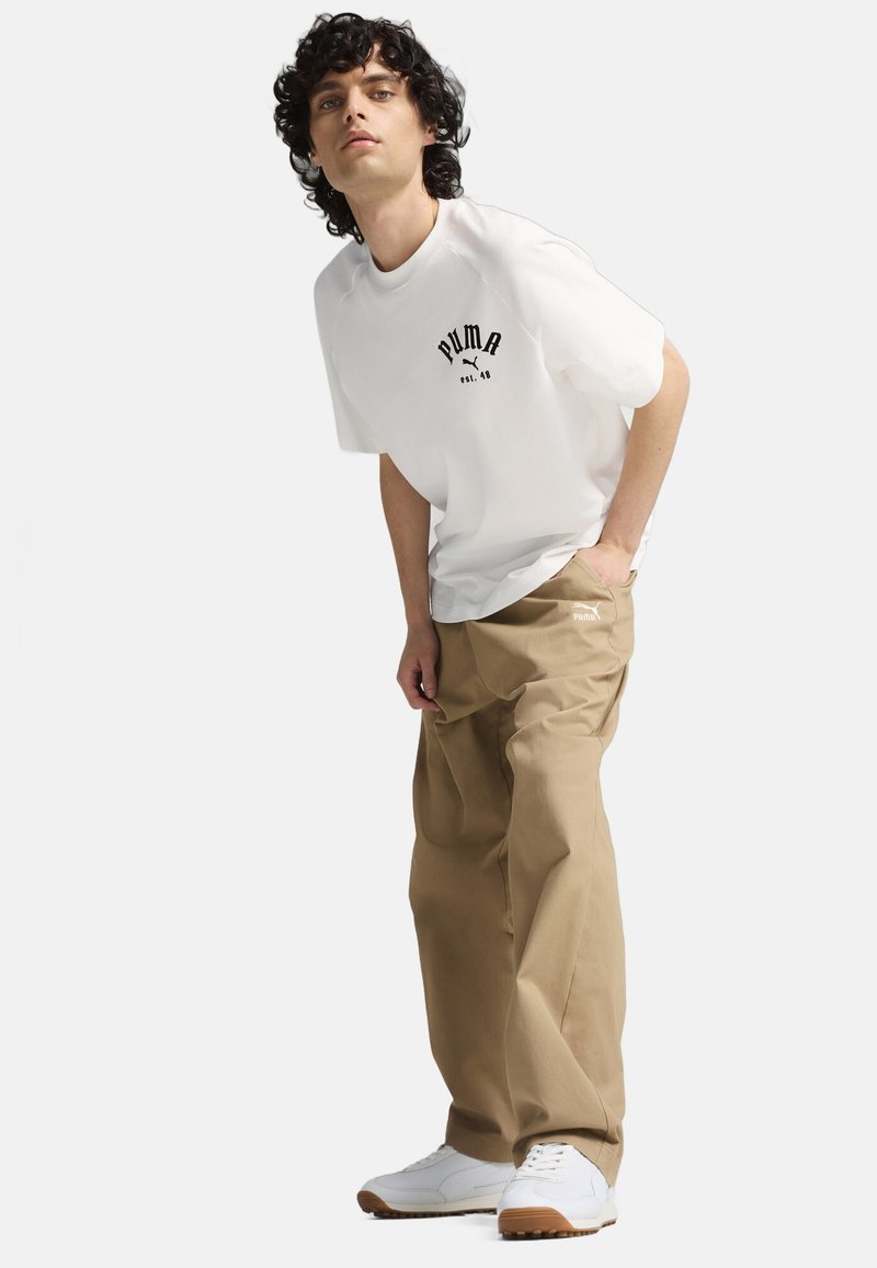 White short-sleeve t-shirt with black "PUMA" logo and "est. 48". Light brown loose-fitting pants. White sneakers with gum soles.