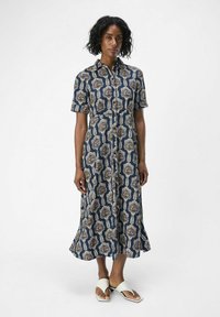 Vestido camisero en azul marino con estampado floral en beige y blanco. Mangas cortas, cuello, cierre frontal de botones y dobladillo acampanado. Llevado con sandalias claras.